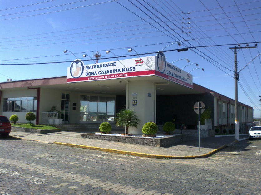 Fachada da Maternidade Dona Catarina Kuss, com uma placa no topo da entrada principal que inclui o logotipo do Governo de Santa Catarina e a inscrição "Secretaria de Estado da Saúde". A edificação tem estrutura térrea, fachada simples com janelas de vidro e canteiros com arbustos podados. Em frente, há uma calçada de pedras irregulares e alguns carros estacionados. O céu está limpo e azul, com diversos cabos de eletricidade cruzando a imagem.