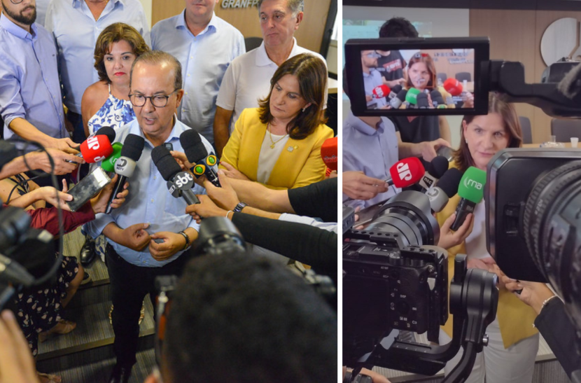 Montagem com duas fotos de uma coletiva de imprensa. À esquerda, um homem de óculos fala à imprensa, cercado por repórteres com microfones de diferentes emissoras, como SBT, ND, CBN e outras. Atrás dele, pessoas observam, incluindo uma mulher de blazer amarelo. À direita, a mesma mulher aparece dando entrevista, com diversos microfones à sua frente. A cena é registrada por uma câmera profissional, cuja tela mostra o rosto da entrevistada em destaque.