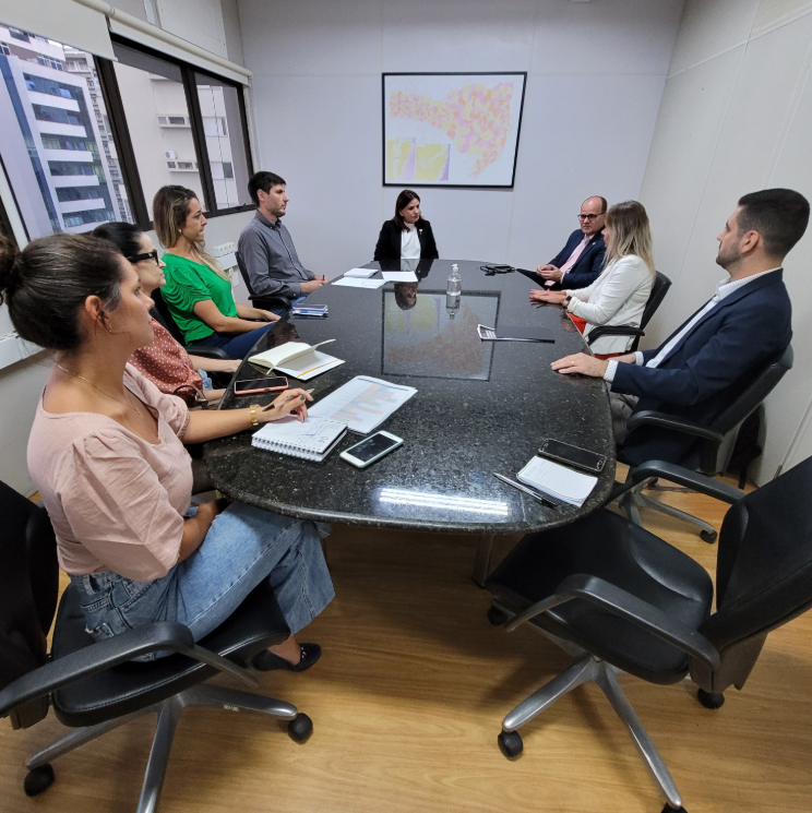 Reunião de trabalho em uma sala corporativa com oito pessoas sentadas ao redor de uma mesa oval de granito. Todos estão atentos e com cadernos, anotações ou celulares à frente. A sala tem janelas amplas com vista para prédios e um quadro colorido na parede ao fundo.