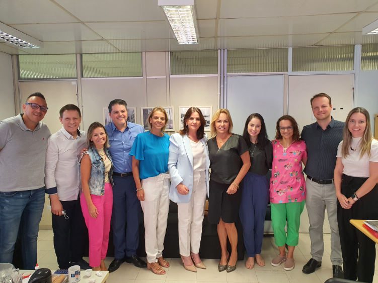 Grupo de 12 pessoas posando para foto em um ambiente de escritório. Todos estão sorrindo e em pé lado a lado, demonstrando clima amigável e profissional. A maioria veste roupas sociais e casuais. A imagem parece ter sido registrada durante uma visita ou reunião institucional.