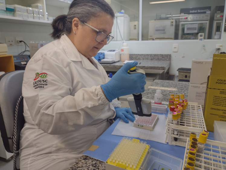 Profissional de laboratório com jaleco branco e luvas azuis manuseia uma micropipeta em bancada de laboratório. Ela trabalha com amostras em tubos de ensaio organizados em racks, e utiliza equipamentos de análise biomédica. O jaleco exibe o logotipo do LACEN/SC (Laboratório Central de Saúde Pública de Santa Catarina). Ao fundo, é possível ver uma bancada com frascos, caixas e equipamentos laboratoriais.