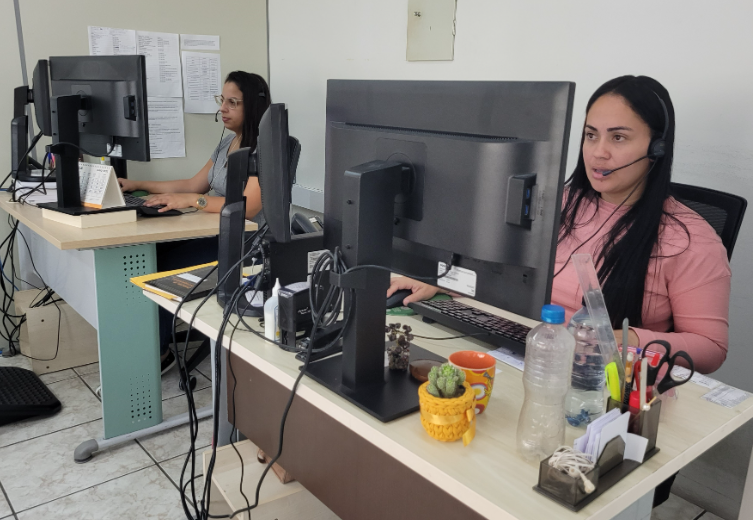 Duas mulheres trabalham em um escritório, sentadas em frente a computadores com fones de ouvido. Elas atendem chamadas, aparentando estar em um serviço de ouvidoria ou atendimento ao público. Há objetos pessoais sobre as mesas, como canecas, garrafa de água, cactos e materiais de escritório.