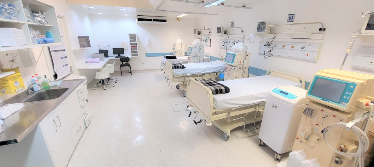 Sala de hemodiálise moderna e bem iluminada, equipada com três leitos hospitalares, cobertores dobrados e máquinas de diálise ao lado de cada cama. No canto esquerdo, há uma pia com bancada e armários contendo materiais hospitalares. Ao fundo, estações de trabalho com computadores e cadeiras brancas completam o ambiente limpo e organizado.