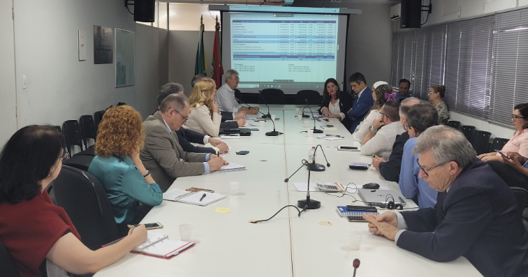 Reunião formal com cerca de 20 pessoas sentadas ao redor de uma longa mesa de reuniões em uma sala corporativa. Todos estão atentos, alguns tomando notas ou utilizando dispositivos eletrônicos. Ao fundo, há uma tela de projeção exibindo uma tabela de dados e duas bandeiras ao lado. O ambiente é bem iluminado, com janelas com persianas fechadas à direita.
