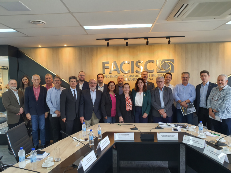 Grupo de aproximadamente 20 pessoas posando para foto em uma sala de reuniões da FACISC (Federação das Associações Empresariais de Santa Catarina), com placa da instituição ao fundo. Na mesa estão garrafas de água, copos, crachás de identificação e materiais de reunião.
