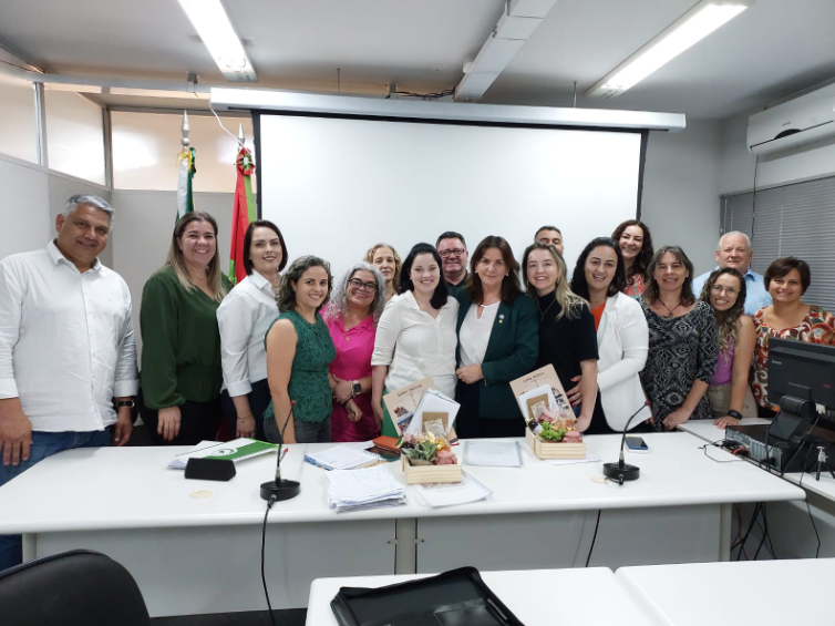Grupo de cerca de 20 pessoas, entre homens e mulheres, posando sorridentes para foto em sala de reunião. No centro da imagem, há duas mulheres segurando folders e cestas de presente. Ao fundo, bandeiras do Brasil e de Santa Catarina e um projetor. Ambiente iluminado, com mesas brancas e equipamentos de escritório.