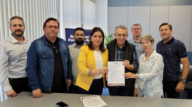 Grupo de oito pessoas posa para foto durante assinatura de documento oficial. Duas pessoas ao centro seguram o papel assinado, em um ambiente interno com mesa, persianas verticais e banner ao fundo com os dizeres 'Regionalização Ponta a Ponta'