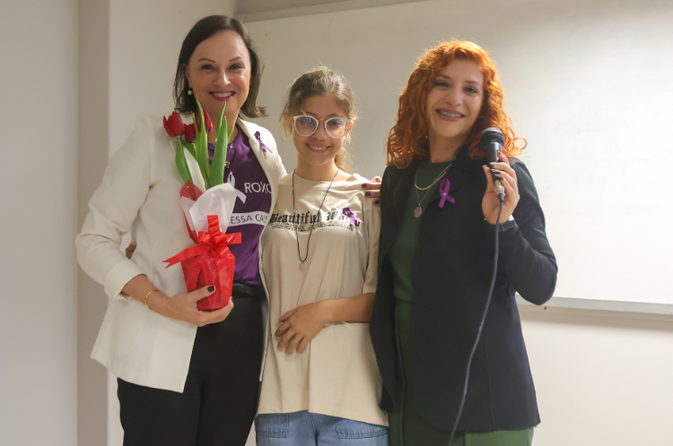 Três mulheres posam sorrindo para a foto. A da esquerda segura um buquê de flores vermelhas, a do centro veste uma camiseta clara com a frase 'Beautiful World' e a da direita segura um microfone. Todas usam laço roxo, símbolo da campanha Maio Roxo de conscientização sobre doenças inflamatórias intestinais.