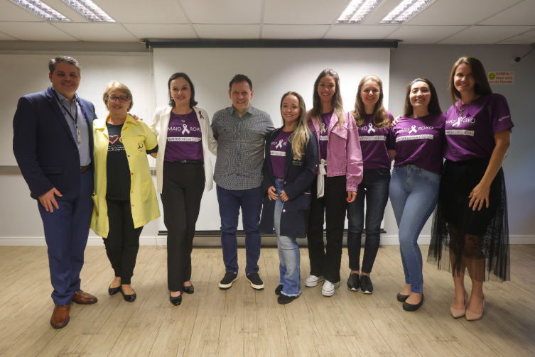 Grupo de nove pessoas posando sorridentes lado a lado em um ambiente interno. A maioria usa camisetas roxas da campanha Maio Roxo, que promove a conscientização sobre doenças inflamatórias intestinais. Ao fundo, um projetor e uma placa na parede com os dizeres: 'Sorria! Você está sendo filmado'.
