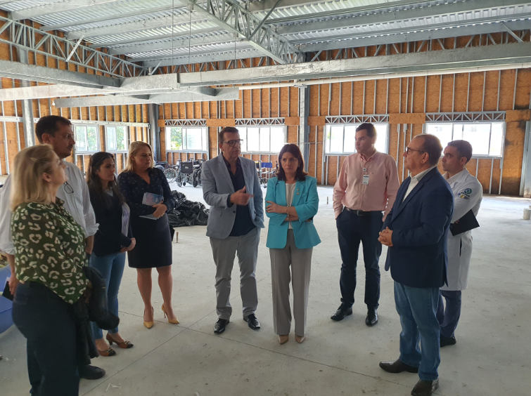 Grupo de profissionais e autoridades em visita técnica a uma obra em andamento de estrutura hospitalar. Estão reunidos em uma sala ampla, ainda em fase de construção, com estrutura metálica aparente e paredes sem acabamento. A mulher no centro, de blazer azul, escuta atentamente um homem que fala gesticulando. O ambiente sugere inspeção ou avaliação do progresso da construção.