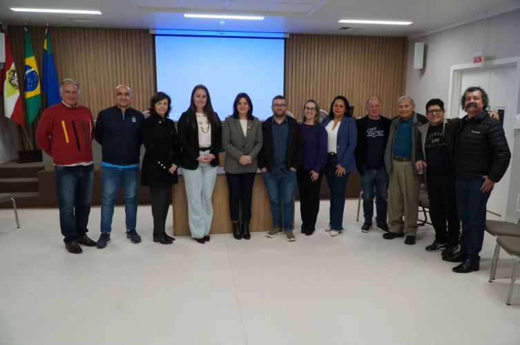 Grupo de pessoas posando para foto em uma sala de reuniões institucional. Estão dispostas em linha à frente de uma mesa e de um telão azul. Ao fundo, aparecem as bandeiras de Santa Catarina, do Brasil e do município. Os participantes, homens e mulheres de diferentes idades, estão sorrindo, demonstrando um momento de confraternização ou finalização de um evento formal.