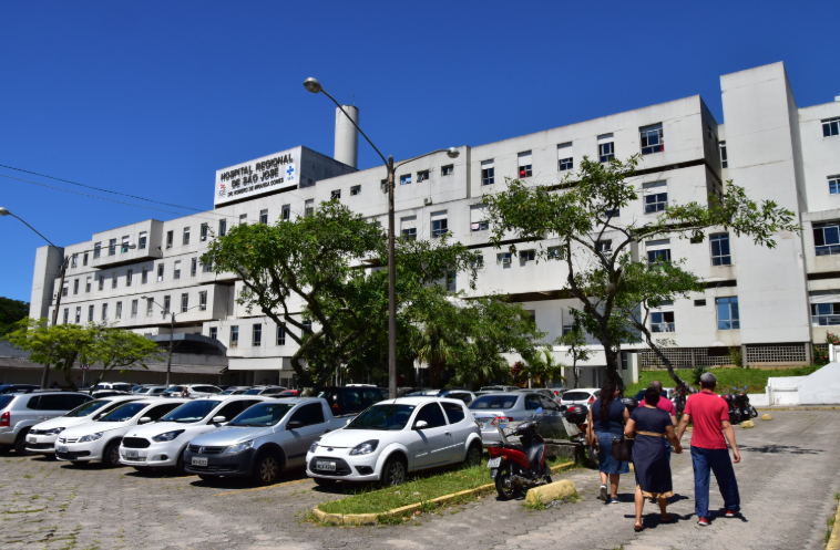 Fachada do Hospital Regional de São José Dr. Homero de Miranda Gomes, com várias janelas, estacionamento cheio de carros e pessoas caminhando em direção à entrada, em um dia ensolarado.