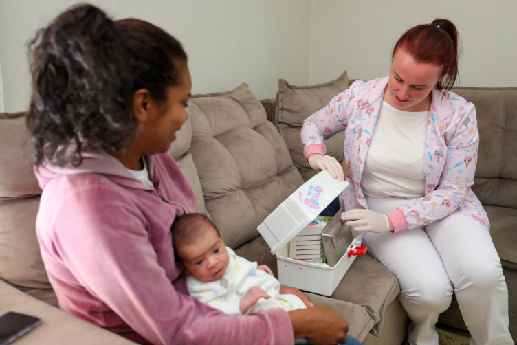 Profissional da saúde com jaleco infantil rosa e luvas brancas prepara kit do “Teste do Pezinho” ao lado de uma mãe com um recém-nascido no colo, sentadas em um sofá durante atendimento domiciliar.