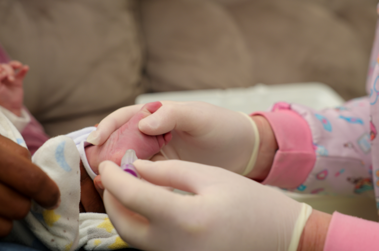 Profissional da saúde usando luvas e jaleco infantil realiza a coleta do “Teste do Pezinho” em um recém-nascido, segurando delicadamente o calcanhar do bebê durante o procedimento.
