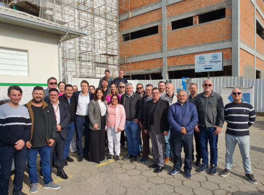 Grupo de cerca de 30 pessoas, incluindo homens e mulheres, posa sorridente em frente a um prédio em construção com estrutura de tijolos expostos. Ao fundo, uma placa indica 'HST - Ambulatório de Saúde - Amigos'. A foto foi tirada em área externa, em dia ensolarado.