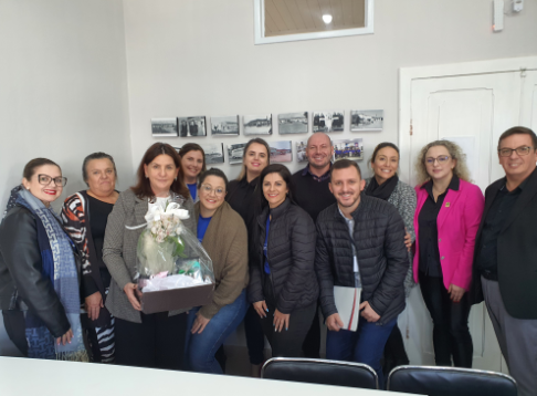 Grupo de 14 pessoas, entre homens e mulheres, posa sorridente em sala com parede branca e fotos históricas em preto e branco. Uma mulher ao centro segura uma cesta de presente com flores. O ambiente é fechado e iluminado, sugerindo ocasião de homenagem ou visita institucional.