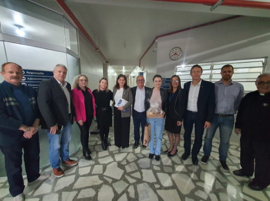 Grupo de 13 pessoas, entre homens e mulheres, posa em corredor de hospital com paredes claras e piso de pedras. Ao centro, uma jovem segura um presente embrulhado. Placas indicam setores como 'Administração' e 'Financeiro'. Relógio de parede marca 10h12. Ambiente fechado e institucional.