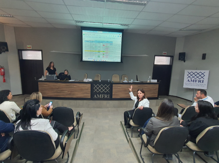 Reunião em auditório da AMFRI, com participantes sentados e uma mulher em destaque falando e gesticulando com a mão levantada. No fundo, uma tela exibe uma planilha colorida com dados. Duas mulheres estão na mesa principal operando o computador. O ambiente é formal e voltado para discussões técnicas sobre gestão pública ou saúde regional. Banner da AMFRI aparece à direita com o nome completo: Associação dos Municípios da Região da Foz do Rio Itajaí.