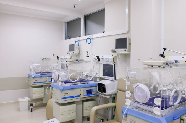 Vista de uma ala neonatal moderna com várias incubadoras infantis alinhadas, cada uma equipada com monitores, sensores e cadeiras para acompanhantes. O ambiente é limpo, bem iluminado e com design funcional, evidenciando tecnologia de ponta e organização hospitalar.