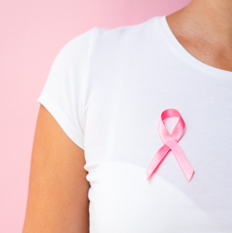 Close no peito de uma pessoa vestindo camiseta branca com uma fita rosa, símbolo da campanha de conscientização sobre o câncer de mama, sobre um fundo rosa.