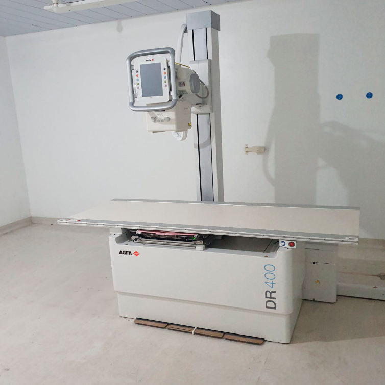Equipamento de radiografia digital da marca AGFA modelo DR 400 em uma sala branca vazia, com piso claro. O aparelho está posicionado com a mesa de exame e o tubo de raio-x vertical. Na parede ao fundo, há duas tomadas azuis e uma pequena estrutura de suporte.