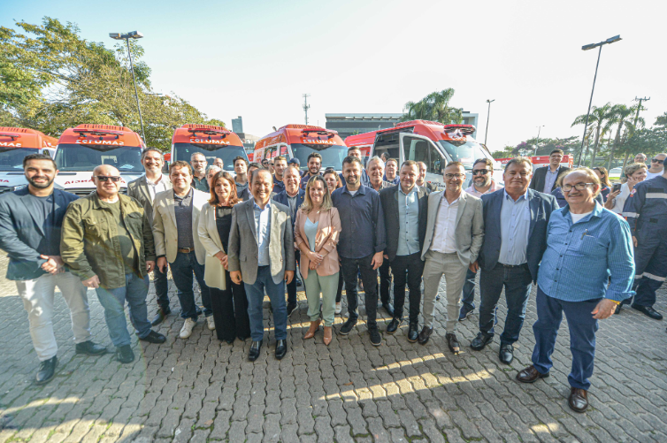 Grupo de cerca de 25 pessoas posando para foto em área externa, em frente a uma fileira de ambulâncias do SAMU 192, estacionadas. As pessoas estão sorrindo, vestindo trajes sociais e casuais, e o clima está ensolarado. Ao fundo, árvores e parte de um prédio são visíveis.