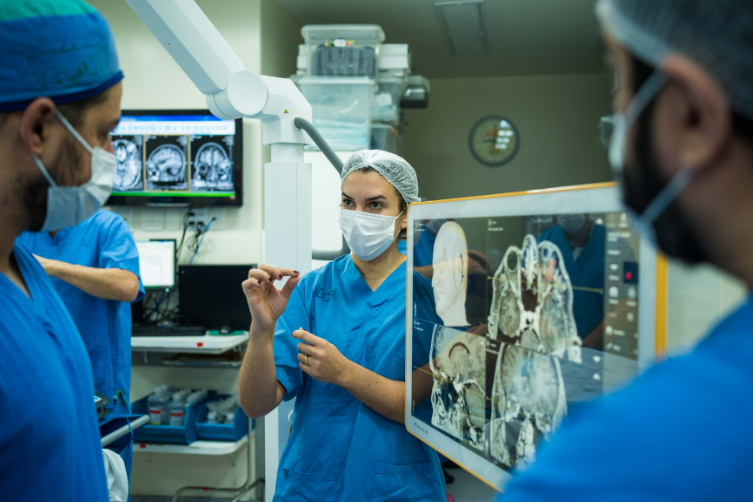 Profissionais da saúde vestidos com roupas cirúrgicas azuis e máscaras discutem exames de imagem em uma sala médica. Uma mulher ao centro gesticula enquanto fala, ao lado de um monitor que exibe tomografias computadorizadas de crânio. No fundo, outros exames são projetados em outra tela.