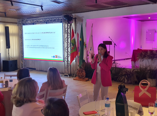 Mulher em pé com microfone, vestindo blazer rosa, apresenta dados sobre o déficit orçamentário da saúde de Santa Catarina em evento. Ao fundo, telão exibe valores e informações financeiras, com bandeiras do Brasil, Santa Catarina e outras. Ambiente iluminado com luz rosa, decoração floral e público sentado em mesas redondas.