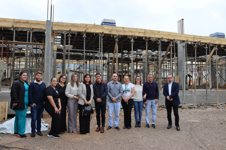 Grupo de profissionais e autoridades posando em frente a uma construção em andamento, com estruturas de concreto e andaimes visíveis ao fundo. A visita ocorre em área de expansão de um hospital ou instituição pública.