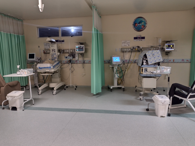 Ambiente hospitalar com duas incubadoras neonatais equipadas com monitores e aparelhos de suporte à vida. As áreas são separadas por cortinas verdes e decoradas com temática infantil, indicando ser uma UTI neonatal.