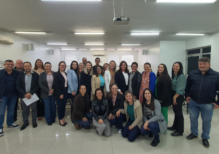 Grupo de gestores e gestoras posando para foto em sala de reuniões, com paredes brancas e iluminação fluorescente.