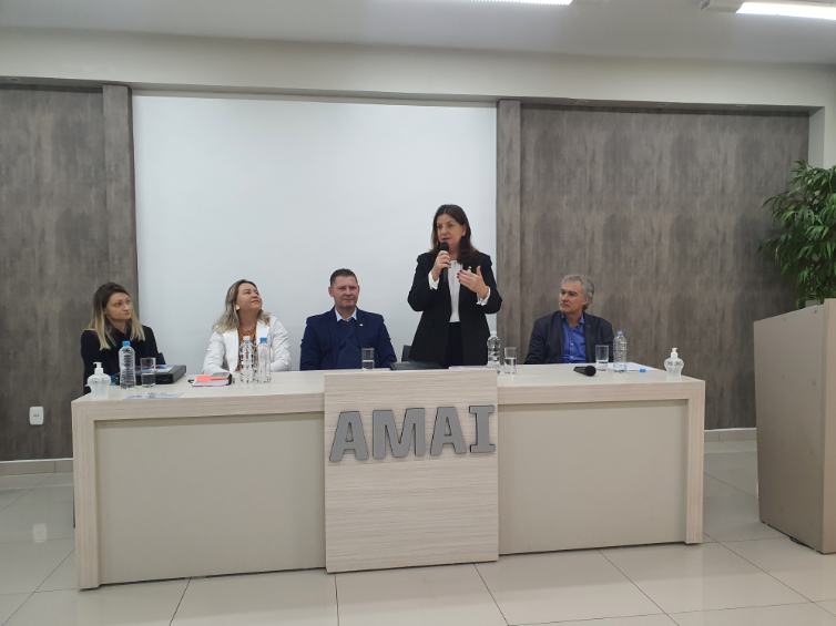 Mulher de terno preto falando ao microfone em evento da AMAI, com quatro pessoas sentadas à mesa e painel ao fundo.