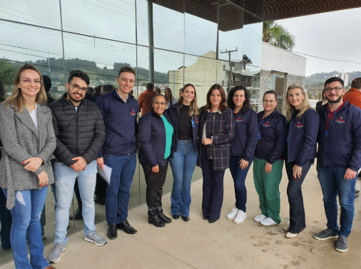 Grupo de 11 pessoas, entre elas profissionais da saúde e uma parlamentar, posando em frente a fachada envidraçada do Hospital de Pinhalzinho.