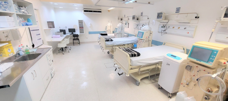 Ambiente hospitalar limpo e moderno, com várias cadeiras hospitalares organizadas ao lado de máquinas de hemodiálise, monitores e equipamentos médicos. Ao fundo, há mesas com computadores e cadeiras de escritório, demonstrando um espaço preparado para atendimento de pacientes renais.