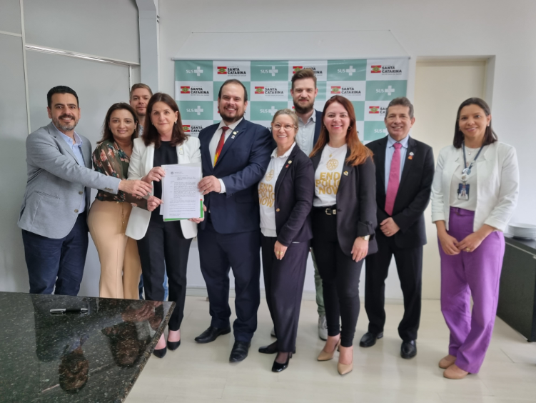 Grupo de pessoas sorrindo e posando para foto em frente a um banner do SUS e do Governo de Santa Catarina. No centro, quatro pessoas seguram um documento oficial, simbolizando a assinatura ou entrega de um acordo ou parceria institucional. Algumas pessoas usam camisetas com a mensagem "END POLIO NOW".
