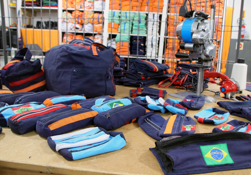 Mesa com estojos, bolsas e mochilas de tecido azul com detalhes coloridos, produzidos em oficina de costura dentro de presídio, com fardos de uniformes empilhados ao fundo.