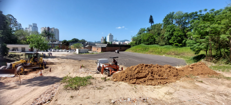 A imagem mostra um amplo espaço aberto em obras, aparentemente sendo preparado para se tornar um estacionamento asfaltado.  Primeiro plano: Há um monte de terra à direita e duas máquinas de construção — uma motoniveladora amarela da marca New Holland e uma retroescavadeira laranja — operando no local.  Centro: O piso de grande parte da área já está nivelado e coberto por asfalto ou brita escura. Algumas pessoas e veículos estão estacionados ao fundo, próximos a um prédio de baixa altura.  Fundo: É possível ver construções urbanas, incluindo prédios altos e áreas arborizadas. À direita, há um talude gramado e árvores densas.  Céu: Azul, com poucas nuvens, indicando um dia ensolarado.