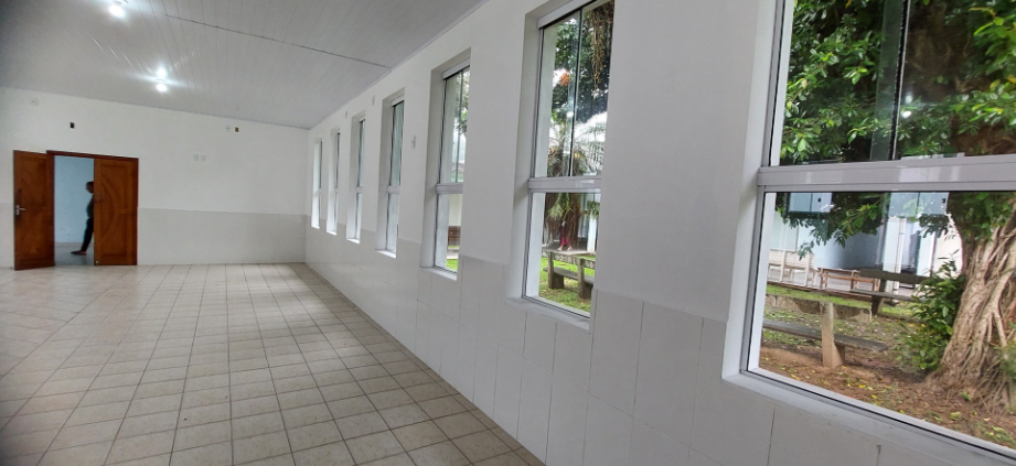 Corredor interno recém-reformado com piso de cerâmica clara, paredes brancas e teto em forro de PVC branco, iluminado por lâmpadas de LED embutidas. À esquerda, portas duplas de madeira abertas, com uma pessoa visível parcialmente no outro cômodo. À direita, uma sequência de janelas retangulares de vidro com esquadrias brancas, permitindo vista para área externa arborizada com bancos de madeira, gramado e troncos de árvores.