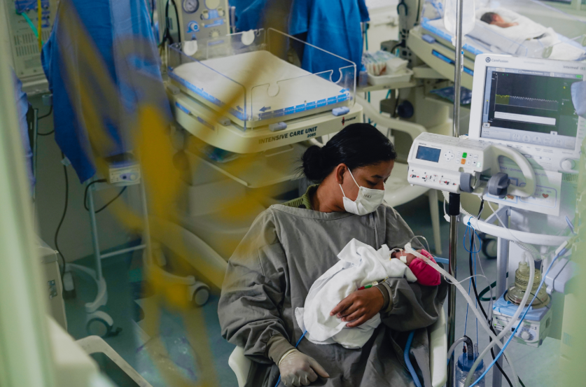 Mulher de pele morena e cabelos pretos presos em coque segura um bebê prematuro nos braços, sentada em uma cadeira ao lado de equipamentos médicos na UTI neonatal. Ela veste máscara facial branca, avental hospitalar cinza e luvas descartáveis. O bebê, enrolado em manta branca e usando gorro rosa, está conectado a tubos de respiração. Ao fundo, há incubadoras e outros recém-nascidos deitados.