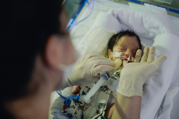Recém-nascido prematuro de pele clara e cabelos pretos, deitado em manta branca dentro de uma incubadora, recebe alimentação por sonda de um profissional de saúde usando luvas descartáveis. O bebê está com os olhos fechados, vestindo roupa estampada infantil, e tem tubos conectados ao nariz para auxílio respiratório. Em primeiro plano, desfocado, aparece parte do rosto do profissional usando máscara.