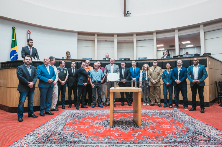 Grupo de autoridades e representantes posa para foto no plenário de uma casa legislativa, posicionados atrás de uma mesa de madeira sobre um tapete vermelho estampado. Ao fundo, há duas bandeiras — do Brasil e de um estado brasileiro — e pessoas sentadas na bancada. Ao centro, um homem segura um documento oficial, cercado por outros participantes em trajes formais e uniformes.