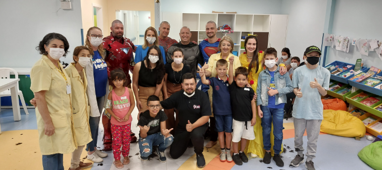 Grupo de voluntários, crianças e profissionais de saúde posam juntos para uma foto em um ambiente infantil com estantes de livros e brinquedos. Alguns adultos estão fantasiados de super-heróis, como Homem de Ferro, Superman, Homem-Aranha, Batman, e de princesa, enquanto outros vestem jalecos ou roupas casuais. Muitas pessoas usam máscaras cirúrgicas, e no chão há mechas de cabelo cortado, sugerindo uma ação de solidariedade ou evento especial.
