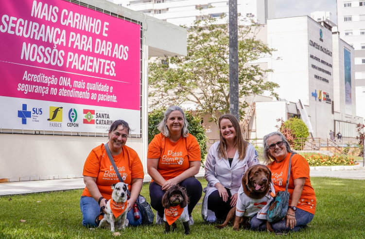 Quatro mulheres agachadas no gramado em frente a um hospital, acompanhadas por três cães vestidos com bandanas ou roupinhas do projeto “Patas Bem do”. Duas mulheres usam camiseta laranja do projeto, uma veste jaleco branco e outra também usa laranja. Ao fundo, há um grande painel rosa com a frase “Mais carinho e segurança para dar aos nossos pacientes” e logotipos do SUS, FAHECE, CEPON e Governo de Santa Catarina.