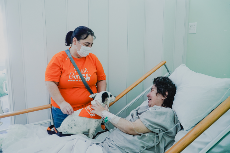 Mulher de camiseta laranja e máscara, integrante do projeto “Patas Bem do”, está ao lado de um leito hospitalar segurando um cachorro pequeno com bandana laranja. O animal está apoiado na cama e interage com uma paciente sorridente, que está deitada com roupas hospitalares e acesso venoso no braço.