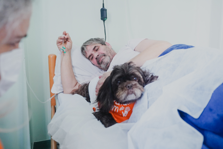 Homem deitado em leito hospitalar, com acesso venoso na mão, acaricia um cachorro pequeno de pelagem preta com bandana laranja do projeto “Patas do Bem”. O animal está deitado ao lado dele sobre o lençol branco, enquanto uma pessoa parcialmente visível à esquerda observa a cena.