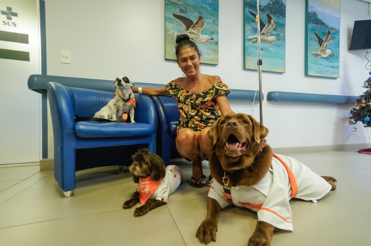 Mulher sorridente, vestindo vestido floral, agacha-se em corredor hospitalar ao lado de três cães do projeto “Patas do Bem”. Um cão pequeno e branco com manchas pretas está sobre uma poltrona azul, usando bandana laranja; um cão pequeno de pelagem marrom-escura está deitado no chão com bandana laranja; e um cão grande marrom, vestido com roupinha branca e laranja, está deitado em primeiro plano com a boca aberta em expressão alegre. Ao fundo, há quadros de aves e uma árvore de Natal.