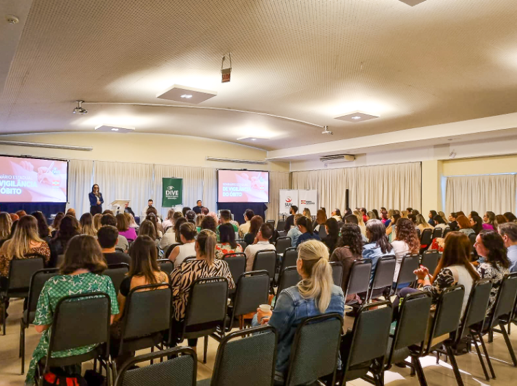 Auditório com várias fileiras de cadeiras ocupadas por participantes assistindo a uma palestra. No palco, quatro pessoas estão posicionadas, sendo uma delas em pé ao lado de um púlpito, falando ao público. Dois telões exibem o título “Seminário Estadual de Vigilância do Óbito” junto ao logotipo da DIVE e do SUS. Atrás, cortinas brancas cobrem a parede e banners institucionais estão posicionados ao fundo.