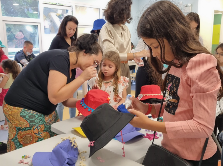 Crianças e adultos participam de uma oficina artesanal em ambiente interno bem iluminado. Sobre as mesas, há chapéus coloridos (vermelho, preto, lilás) sendo decorados com pompons, fitas e outros enfeites. Uma menina de blusa rosa segura um tubo de cola enquanto customiza um chapéu preto. Ao fundo, outras pessoas também trabalham na decoração, interagindo e ajudando as crianças.