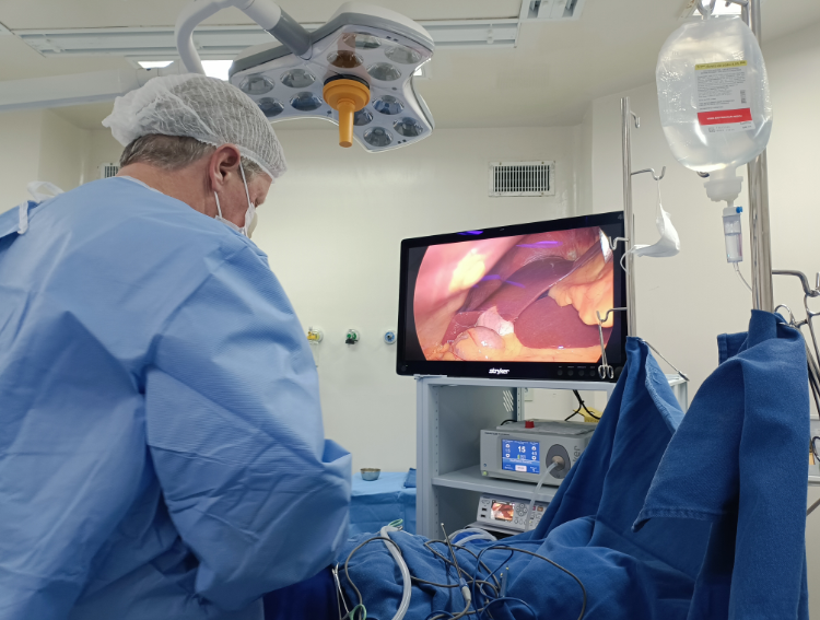 A imagem mostra um cenário de cirurgia laparoscópica. Um profissional de saúde, vestido com avental cirúrgico azul, touca e máscara, está posicionado à esquerda, operando o paciente. Na frente dele, há um monitor exibindo imagens internas do corpo, mostrando órgãos e tecidos, provavelmente capturados por uma câmera laparoscópica.  Ao lado do monitor, vê-se um suporte com soro fisiológico pendurado e equipamentos médicos como cabos e dispositivos eletrônicos para monitoramento e controle da cirurgia. O ambiente é claro, com iluminação cirúrgica no teto, característico de um centro cirúrgico.