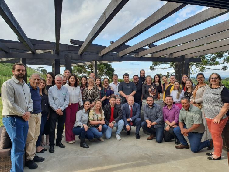 A imagem mostra um grupo numeroso de pessoas posando para uma foto coletiva ao ar livre, sob uma estrutura de madeira com vigas horizontais, que lembra um pergolado.  O cenário ao fundo inclui vegetação verde e montanhas, sugerindo um ambiente serrano. O grupo é composto por homens e mulheres de diferentes idades, alguns em pé e outros agachados ou ajoelhados na frente. A maioria está sorrindo e olhando para a câmera, transmitindo um clima amistoso e descontraído, embora com um toque de formalidade — algumas pessoas usam roupas sociais, como camisas, blazers e gravatas, enquanto outras estão com trajes mais casuais.  A luz natural e o céu parcialmente nublado completam a atmosfera de encontro profissional ou institucional em um local aberto e agradável.